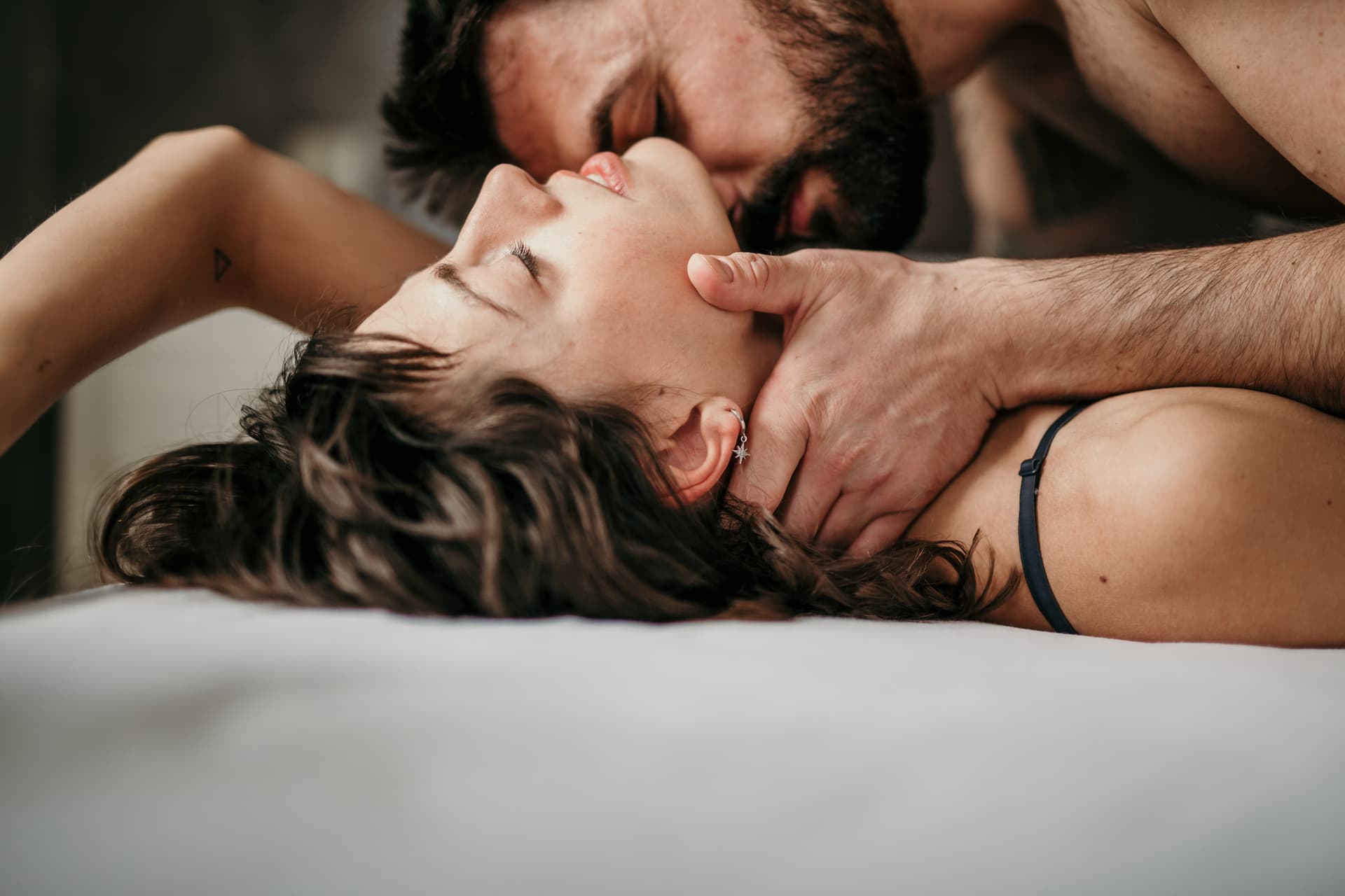 Side view photo of a beautiful couple in bed, being gentle and sensual. Handsome guy with a beard on top of the beautiful brunette is kissing her neck while her eyes are closed. Making love concept